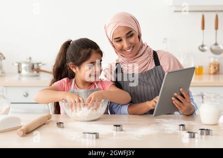 Madre musulmana che cuoce con la figlia piccola a casa, che verifica i biscotti Ricetta in linea sul tablet digitale, mamma islamica felice in Hijab che gode di cottura Homem Foto Stock