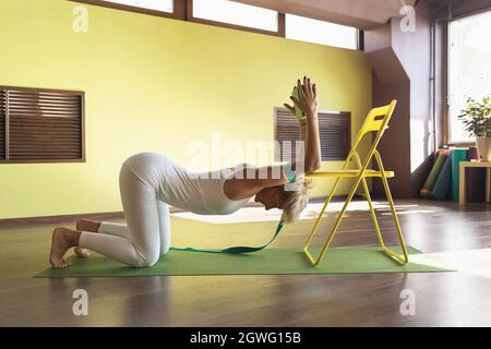 Donna che pratica la terapia yoga, esegue un esercizio per tendere i muscoli della spina dorsale utilizzando una sedia e cinture, treni in studio su un tappeto Foto Stock