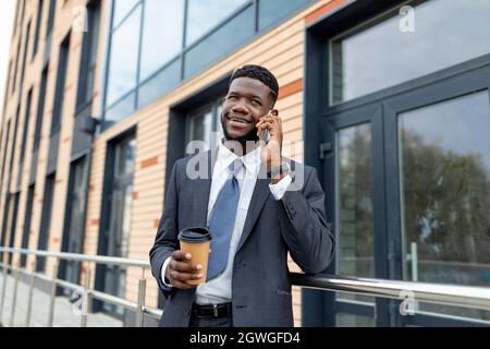 Uomo d'affari nero che parla con il cliente durante la pausa, camminando in città e bevendo caffè, spazio libero. Uomo felice che tiene il caffè per andare e parlare o Foto Stock