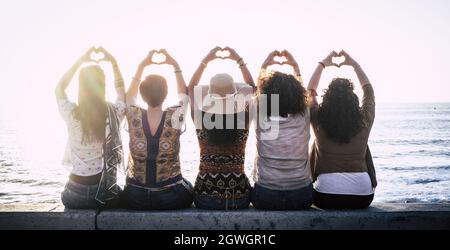 Vista posteriore di cinque donne che si siedono in fila sul muro di mare gesturando l'amore facendo il simbolo della forma del cuore con le mani e ammirando il bel mare. Retro di f Foto Stock