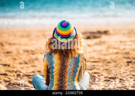 Vista posteriore della donna solitaria seduta sulla sabbia in spiaggia. Donna che si rilassa tranquillamente mentre si siede sulla spiaggia. Schiena di donna in cappello ammirando bella stagcape Foto Stock