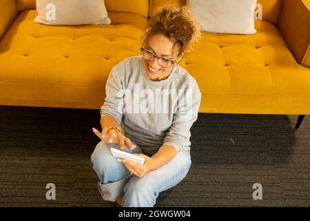 Giovane donna seduta sul pavimento e con tablet digitale. Donna caucasica che controlla i contenuti dei social media sul tablet digitale. Donna felice che naviga in internet Foto Stock
