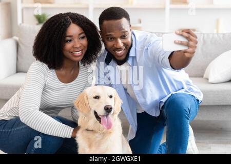 Bella coppia afroamericana in posa e prendendo selfie con cane sul telefono cellulare mentre si riposa a casa e seduto sul pavimento, fuoco selettivo. H Foto Stock