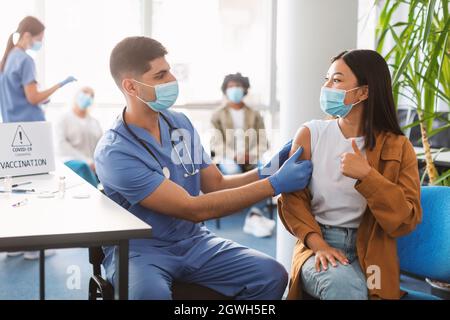 Giovane donna in maschera protettiva per il viso che mostra il gesto dei pollici dopo aver ricevuto l'iniezione in ospedale. Medico che applica la bendatura adesiva sulla donna ' Foto Stock