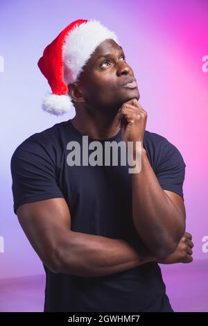 Ritratto di un uomo afro-americano in santa Hat e T-shirt casual su sfondo colorato, ora di natale. Vacanze invernali e persone emozionali Foto Stock
