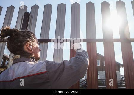 Un uomo dipinge una recinzione in ferro nel giardino e ascolta la musica in cuffia. Foto Stock