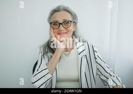 Donna d'affari grigia senior con capelli in giacca a righe con occhiali sta lavorando nel suo ufficio seduto alla scrivania. Buon pensionamento, occupazione e. Foto Stock