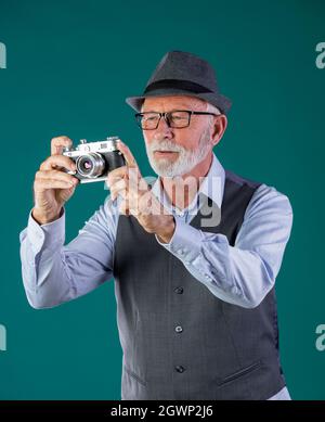 Uomo anziano con barba bianca, in abiti d'epoca e cappello che tiene la vecchia telecamera analogica su sfondo blu in studio Foto Stock