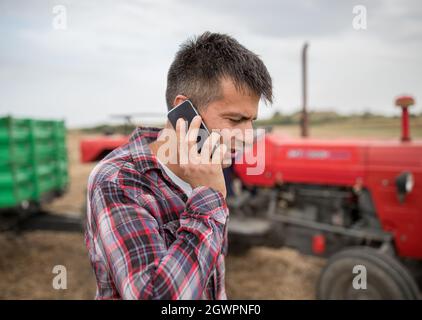 Giovane agricoltore attrattivo che parla al telefono mentre si trova all'aperto in un campo di raccolta davanti al trattore con rimorchio sullo sfondo. Foto Stock