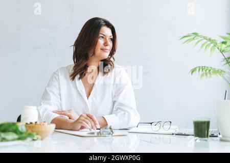 Ritratto di giovane bruna sorridente donna dottore nutrizionista più formato in camicia bianca che lavora al computer portatile nella moderna e luminosa sala d'ufficio. Il medico Presc Foto Stock