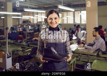 Ritratto di una donna felice in una fabbrica di scarpe che tiene e mostra nuovi uomini scarpe in pelle. Foto Stock
