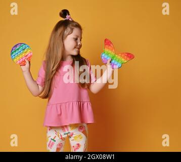 La ragazza del capretto in vestiti rosa mostra due giocattoli sensoriali di colore dell'arcobaleno - lo schioccano. Tiene rotondi e guarda la forma della farfalla uno Foto Stock