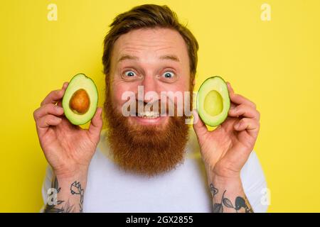 L'uomo felice con barba e tatuaggi tiene un avocado Foto Stock