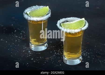 Due colpi di tequila d'oro, con bordi di sale e fette di lime, su sfondo nero Foto Stock