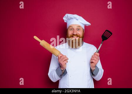 lo chef felice con barba e grembiule rosso tiene la spina di legno Foto Stock