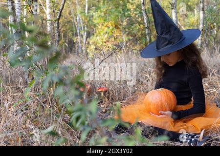 Bambina strega con zucca in mani, seduta in una foresta. Concetto di Halloween. Foto Stock