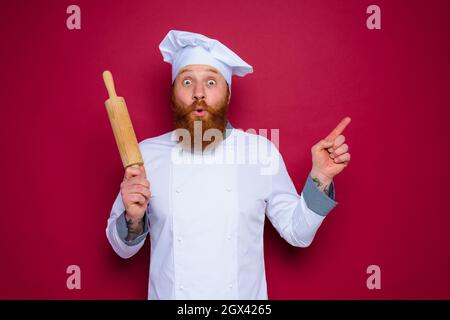 chef stupito con barba e grembiule rosso chef tiene in legno rolling pin Foto Stock