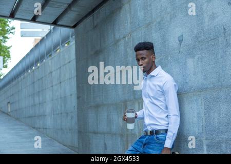 giovane uomo d'affari nero appoggiato a parete con bicchiere di caffè e valigetta in mano Foto Stock