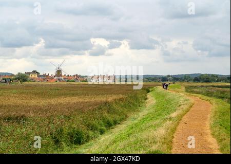 Persone che camminano sul sentiero circolare Blakeney Freshes con vista a Cley sul villaggio di mare con il suo mulino a vento, Norfolk, Inghilterra. Foto Stock