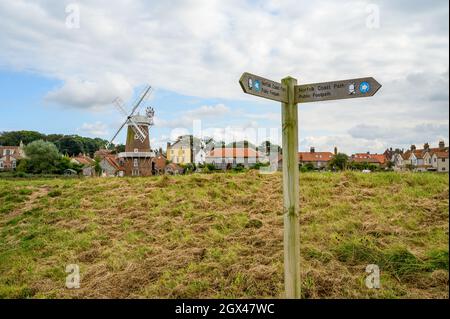 Un cartello di legno per le indicazioni per Norfolk Coast Path sentiero pubblico con Cley sul mare e mulino a vento sullo sfondo, Norfolk, Inghilterra. Foto Stock