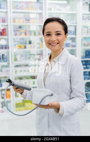 cheerful farmacista in cappotto bianco che tiene tonometro digitale in farmacia Foto Stock
