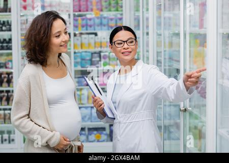 farmacista asiatico in occhiali che tengono appunti e puntano agli scaffali vicino a donna incinta allegra in farmacia Foto Stock