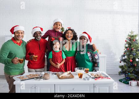 allegra famiglia afroamericana in babbo natale cappelli guardando la macchina fotografica mentre si preparano i biscotti di natale in cucina Foto Stock