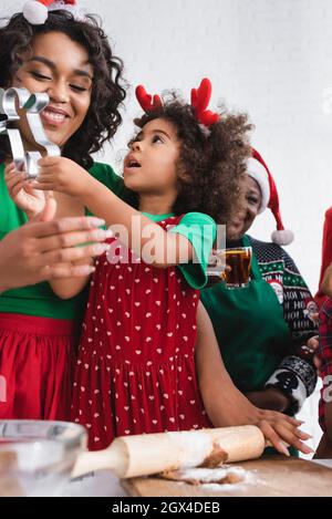 ragazza africana americana in corna di renna fascia che tiene taglierina biscotto vicino mamma felice e sorridente nonna Foto Stock