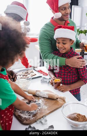 uomo afroamericano con tè alla cannella d'arancia che abbraccia il figlio eccitato vicino alla famiglia che prepara biscotti di natale Foto Stock
