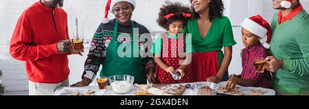 happy afroamericana donne e bambini che preparano biscotti di natale vicino agli uomini che bevono tè alla cannella arancione, banner Foto Stock