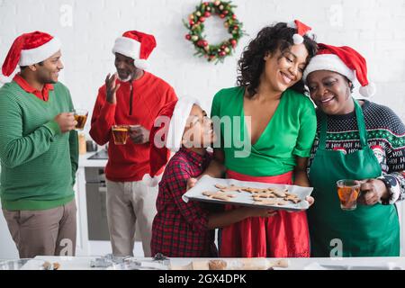 donna afroamericana che tiene la teglia da forno con i biscotti di natale vicino famiglia felice in cucina Foto Stock