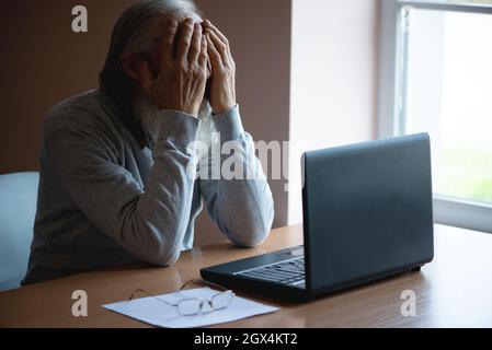 Un anziano stanco, rotto, rassegnato, preoccupato si siede davanti ad un computer portatile e gli copre il volto con la mano. Il triste anziano controlla il suo ac Foto Stock
