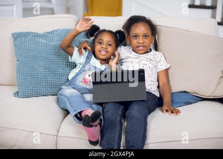 Abbastanza divertente due sorelle afroamericane sorridenti e agitando le mani sulla macchina fotografica seduta sul divano accogliente a casa con il tablet digitale. Concetto di buona infanzia e tecnologia moderna. Foto Stock