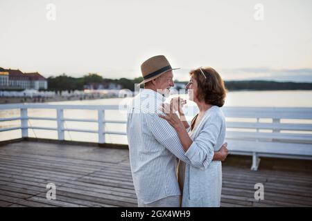 Coppia anziana felice in amore ballando all'aperto sul molo dal mare, guardarsi l'un l'altro. Foto Stock