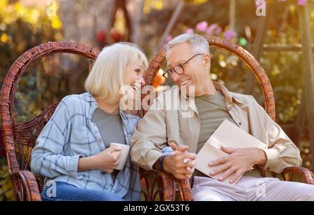 Coppia senior romantica seduta in giardino in sedie di vimini, guardando eah altro con amore e godendo calde giornate autunnali Foto Stock