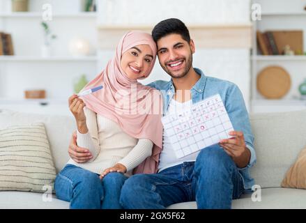 Amorevole coppia islamica tenendo positivo gravidanza test e ovulazione calendario, seduto sul divano e sorridendo alla macchina fotografica Foto Stock