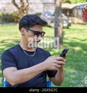 Uomo latino con barba corta e occhiali da sole in profilo seduto nel parco chiacchierando con smartphone cellulare. Verticale Foto Stock