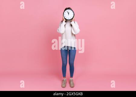 Ritratto a tutta lunghezza di donna sconosciuta che nasconde il suo volto dietro il grande orologio da parete, mostrando il tempo di andare, indossando bianco stile casual maglione. Studio interno girato isolato su sfondo rosa. Foto Stock