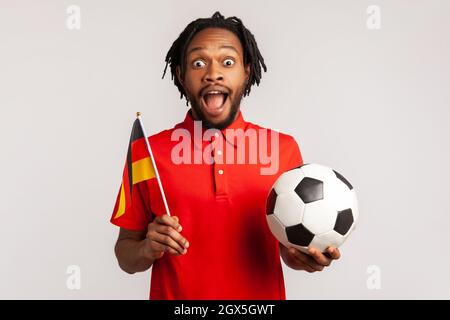 L'uomo stupito con i dreadlock che indossano una T-shirt rossa casual, che sostiene la squadra di calcio tedesca in campionato, allegria e saluto, patriottismo. Studio interno girato isolato su sfondo grigio. Foto Stock