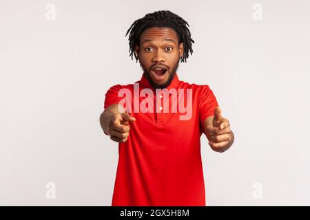 Uomo stupito con dadlock che indossa una T-shirt rossa casual, punta il dito verso la fotocamera, guardando con sorpresa, notando e facendo la scelta. Studio interno girato isolato su sfondo grigio. Foto Stock