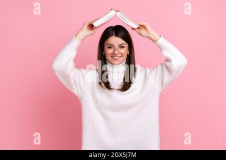 Buona donna osservante in vetri che tengono il libro aperto sopra la testa, esprimente positivo, istruzione, indossando il maglione di stile casual bianco. Studio interno girato isolato su sfondo rosa. Foto Stock