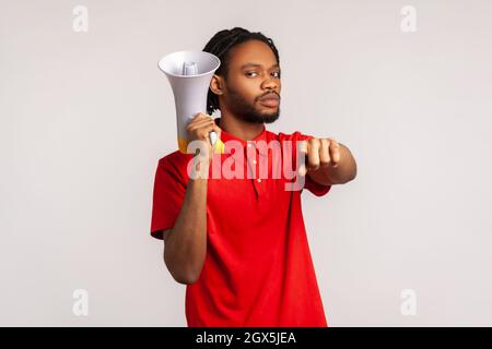 Uomo serio e rigoroso con dreadlock che indossano una T-shirt rossa casual, puntando il dito sulla fotocamera tenendo l'altoparlante in mano, parlando con te, protestando. Studio interno girato isolato su sfondo grigio. Foto Stock