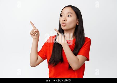 Primo piano ritratto di donna asiatica sorpresa che punta e guarda a sinistra lo spazio vuoto, mostrando il logo, essere impressionato da notizie di vendita, sfondo bianco Foto Stock