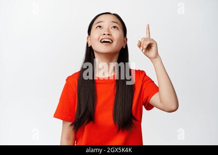 Primo piano della ragazza asiatica stupita guardando qualcosa di impressionante, puntando il dito e guardando in su con il volto sorridente impressionato, in piedi in t-shirt rossa sopra Foto Stock