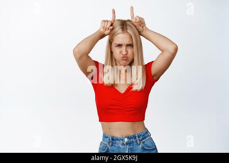 La ragazza bionda di Sassy mostra il gesto dei tori del corno, essendo testardo, pouting e fissando serio alla macchina fotografica, sfondo bianco Foto Stock