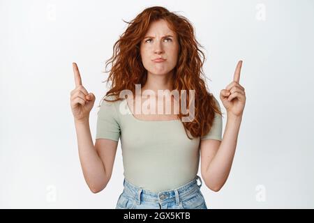 Immagine della ragazza rossa che urlava, punta le dita verso l'alto e guarda turbata, arrabbiata per qualcosa, in piedi in t-shirt su sfondo bianco Foto Stock