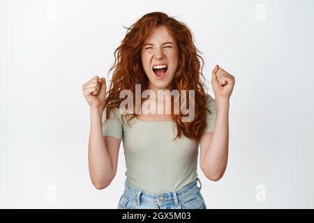 La ragazza rossa eccitata urla, scuote le mani e celebra la vittoria, trionfa e gridando, urla, urla per incoraggiarti, in piedi su bianco Foto Stock