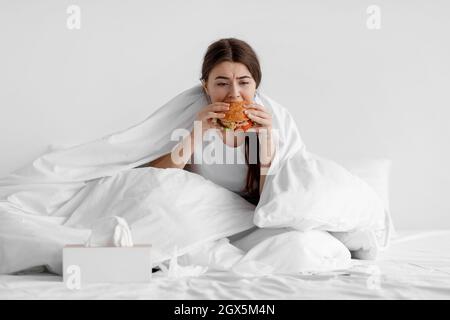 La giovane donna europea affamata si siede a letto a casa e mangia hamburger Foto Stock