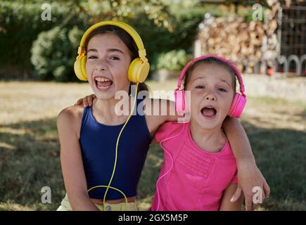 Vista frontale di due ragazze sorridenti e ridenti davanti alla fotocamera con le cuffie sopra la testa. Due ragazze ridono davanti alla macchina fotografica mentre ascoltano Foto Stock