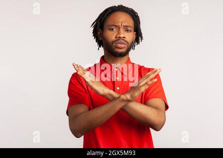 Ritratto di giovane uomo adulto portatore con dreadlock indossare T-shirt rossa, mani incrociate, avvertimento gesturante o proibizionismo, che significa fine corsa. Studio interno girato isolato su sfondo grigio. Foto Stock